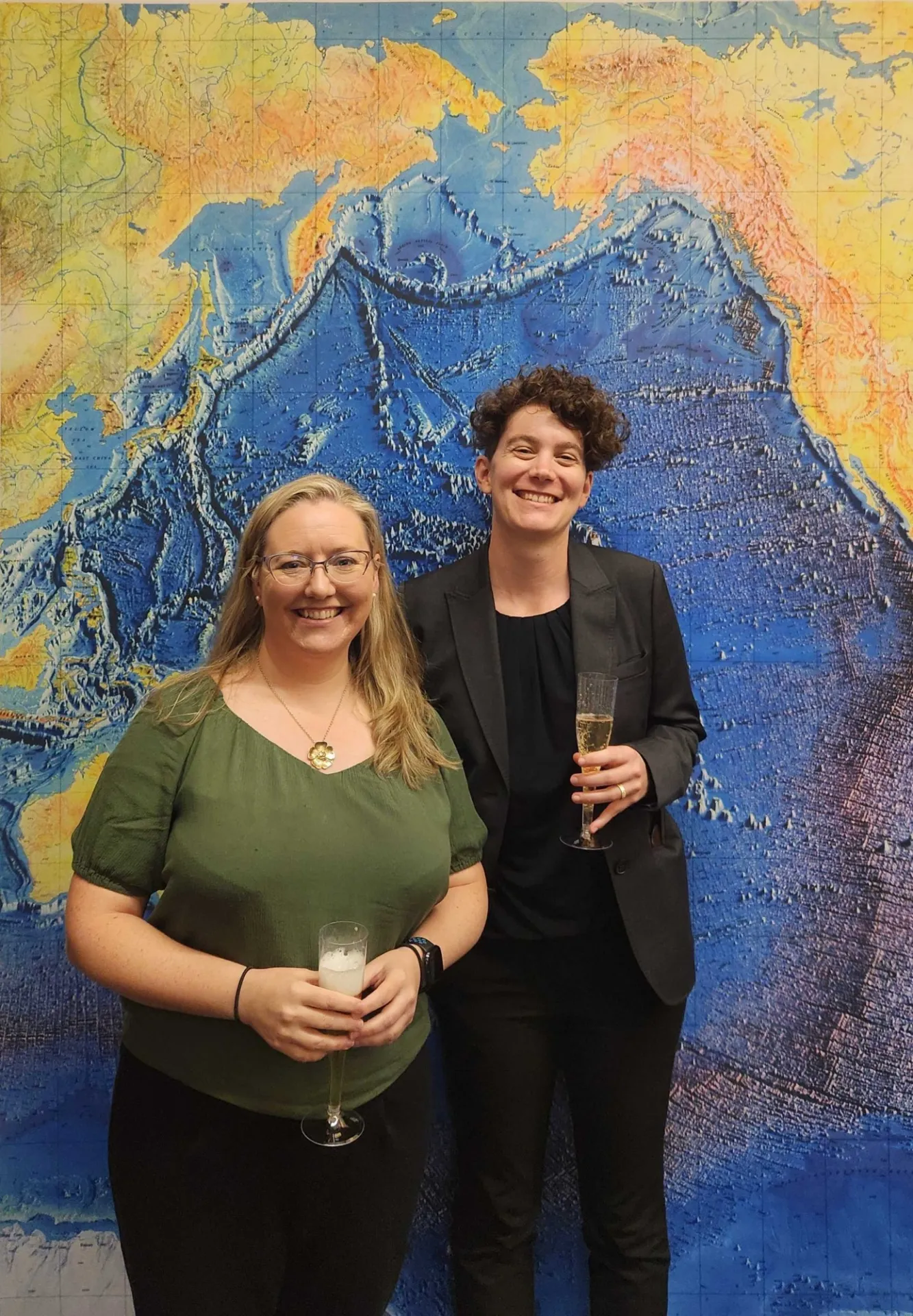 Roisin and Ludda standing in front of a global map with Ludda's focus area, Alaska, shown above them. 